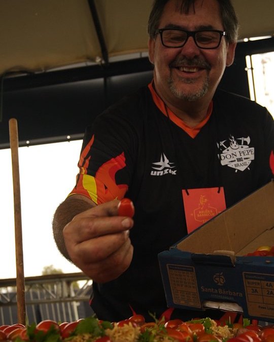 Don Pepe sorrindo e oferecendo um tomate assado na brasa